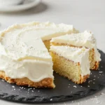 Eine Torta Raffaello auf einer Tortenplatte, mit heller Kokoscreme, weißen Kokosraspeln und dekorativen Pralinen auf der Oberfläche.